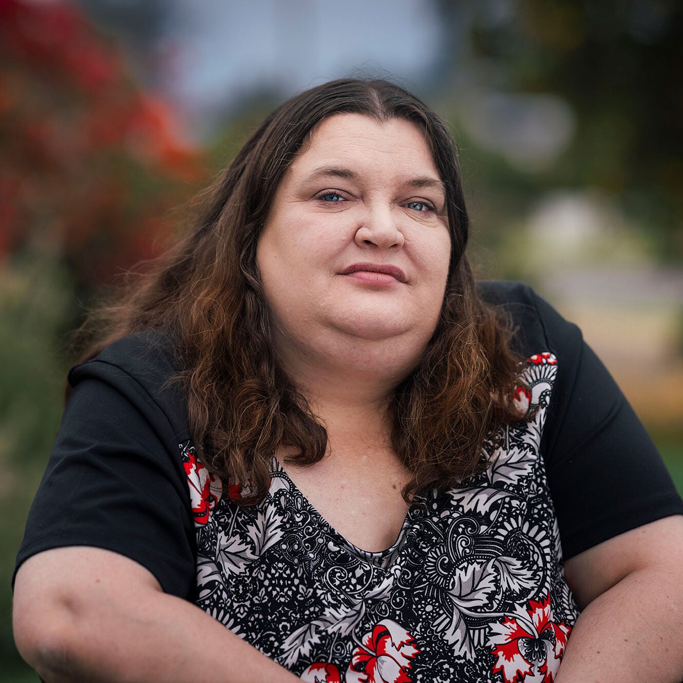 Mel is seated outdoors, looking directly at the viewer with a somber expression. The background is blurred greens and reds.