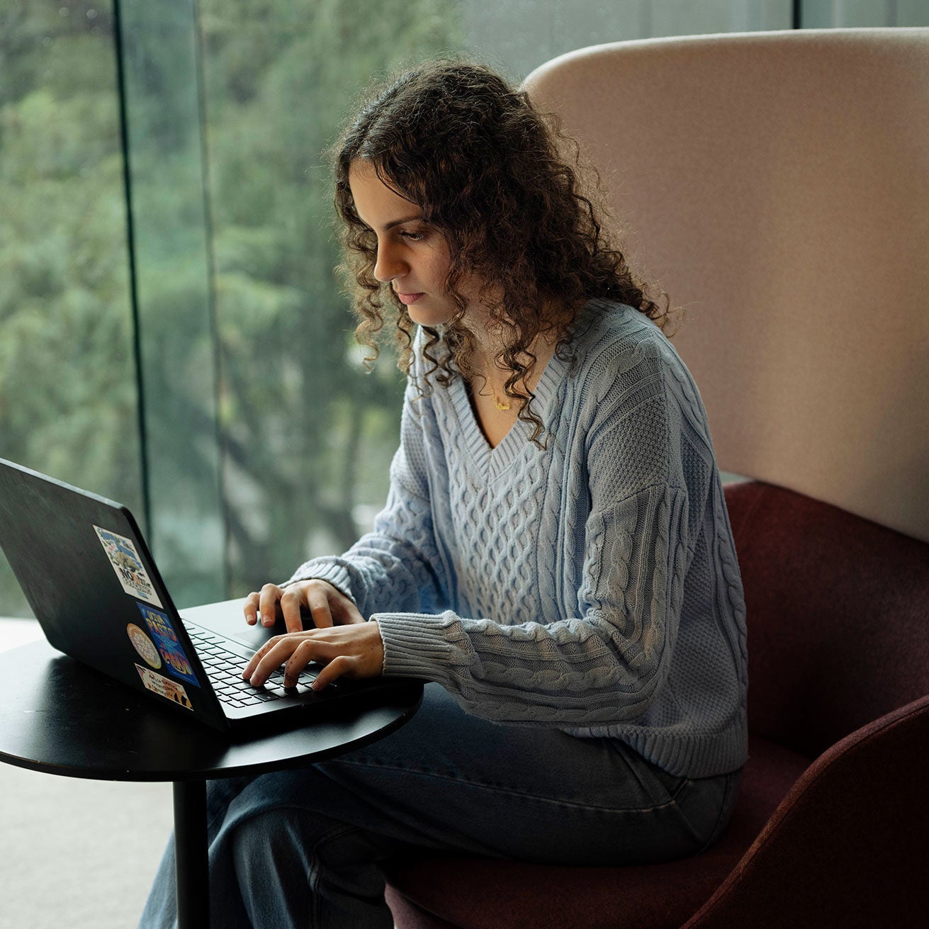Sama sits in a high-backed chair at a small circular desk, looking down at a laptop on the desk as she types. There are four large stickers neatly placed stuck on the top of the laptop, decorating it. Sama sits with her back to the floor-to-ceiling glass window that overlooks a green, out-of-focus landscape.