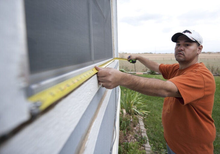 A homeowner stretches out measuring tape along the bottom of a window frame outside of their home.