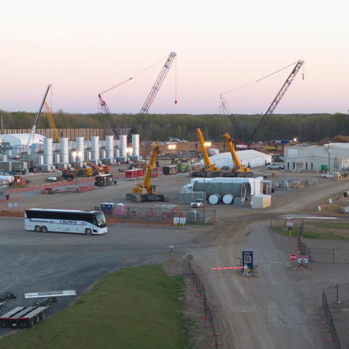 In a large open area surrounded by dense forest, a row of gas-power turbines are flanked by three tall cranes, and construction and other industrial equipment. Dirt roads lead into the area.