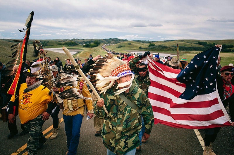 A group of protestors march on a road. In front, leading the group is Catcher Cuts The Rope. He is wearing a camouflage print jacket, a feather headdress, and carrying a swagger stick. To the right, other protestors are holding up a flag of the Unites Sta