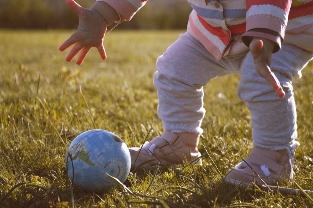 A toddler stands in a grassy field, reaching down with open hands to grasp a small ball in the design of the globe.