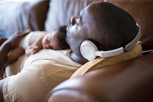 A father and newborn baby daughter at home sleeping. He is wearing headphones and wearing casual clothing.
