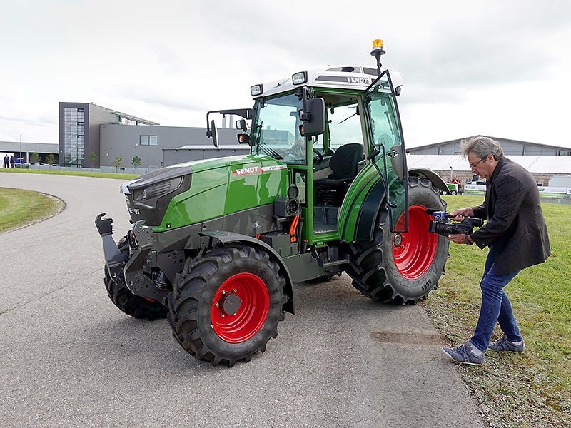 A camera man looks down at camera filming an electric, green tractor.  In its mission to electrify everything, California is considering regulations for zero emissions off-road vehicles like tractors.