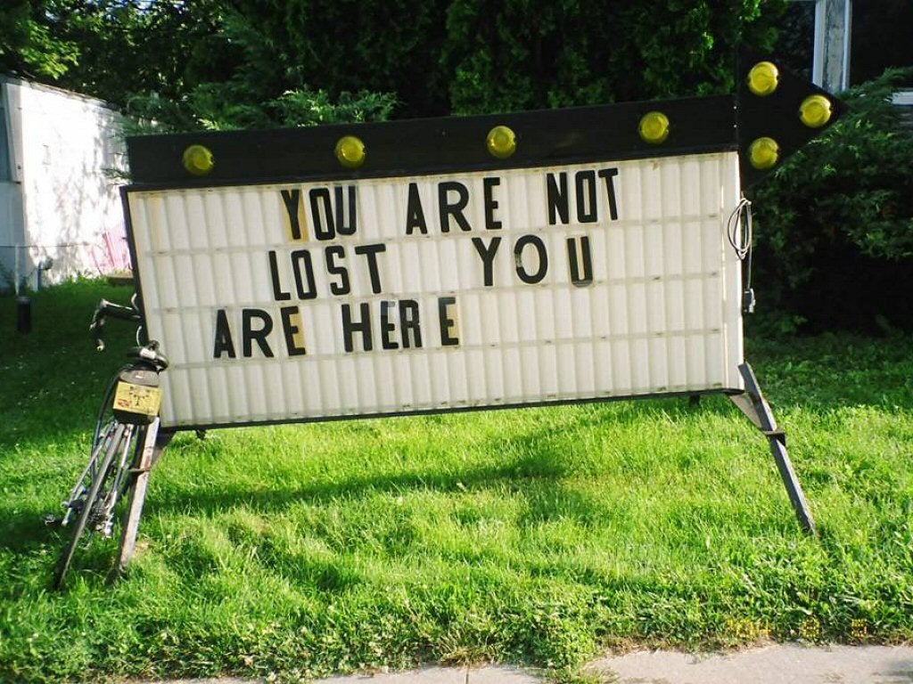 A bicycle is leaning against an antique-style marquee letterbox sign on the grass that reads, "You are not lost. You are here."