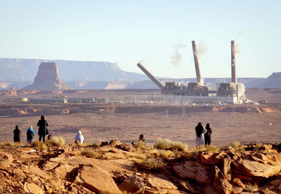 The three 775-foot stacks of Navajo Generating Station outside of Page, Ariz., are imploded on the morning of December 18, 2020. Backs of observers watching from across the way from an elevated landscape.