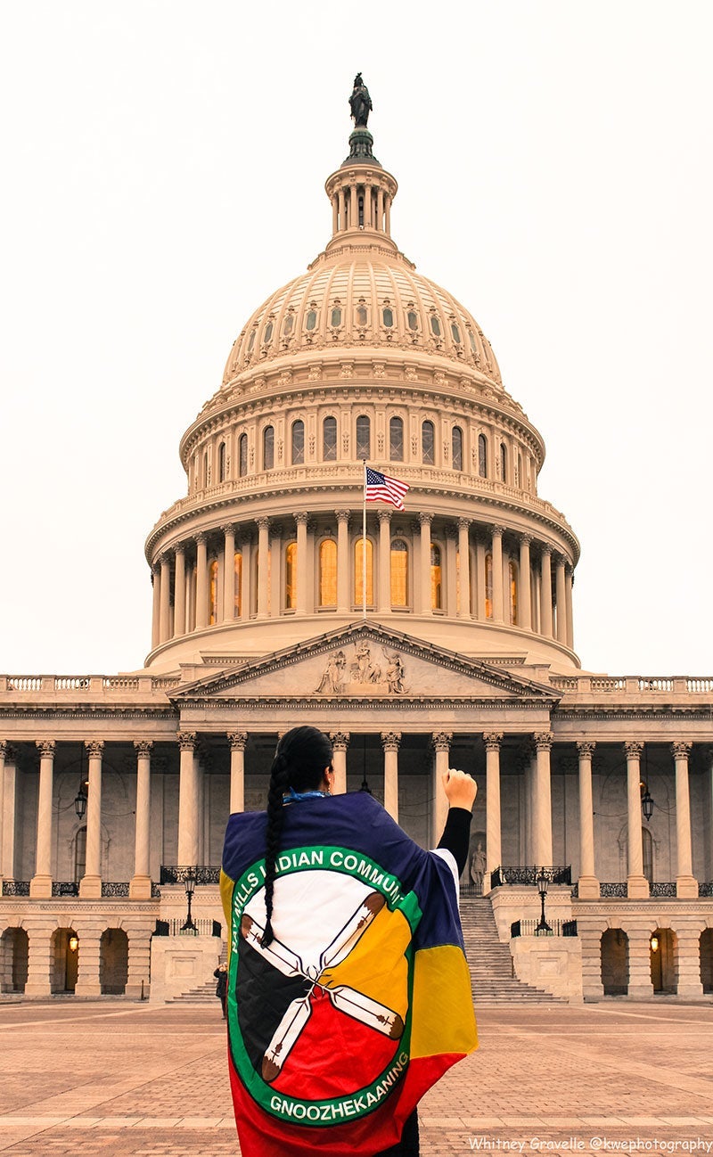 Whitney Gravelle standing in front of the Capitol building at the Women’s March protest in Washington, D.C., in January of 2017. Bay Mills Indian Community flag wrapped around her back, and right fist raised to the air.